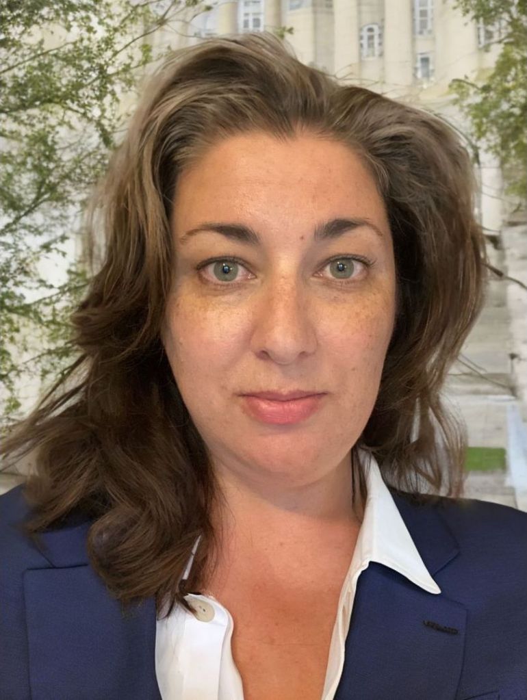 A headshot of Jennifer LaRue; she has brown hair and greyish green eyes. She is wearing a blue blazer and a white blouse. She is standing in front of a white academic building. 